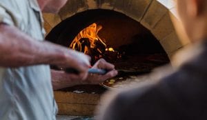 A man using a pizza peel, putting pizza inside a woodfire pizza oven