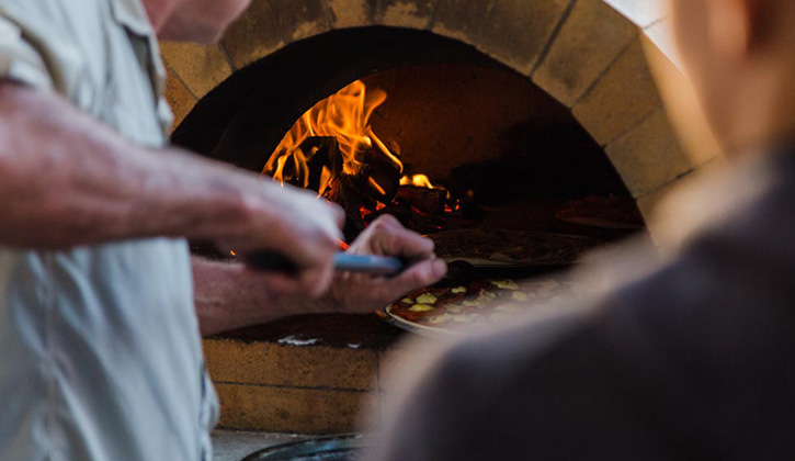 Man placing a pizza into a woodfire oven in a Shellharbour Caravan park